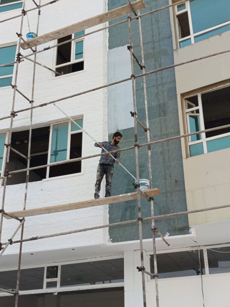 Illustrative library photograph, not an Elbaz project: a worker on a scaffold plank painting an exterior wall
