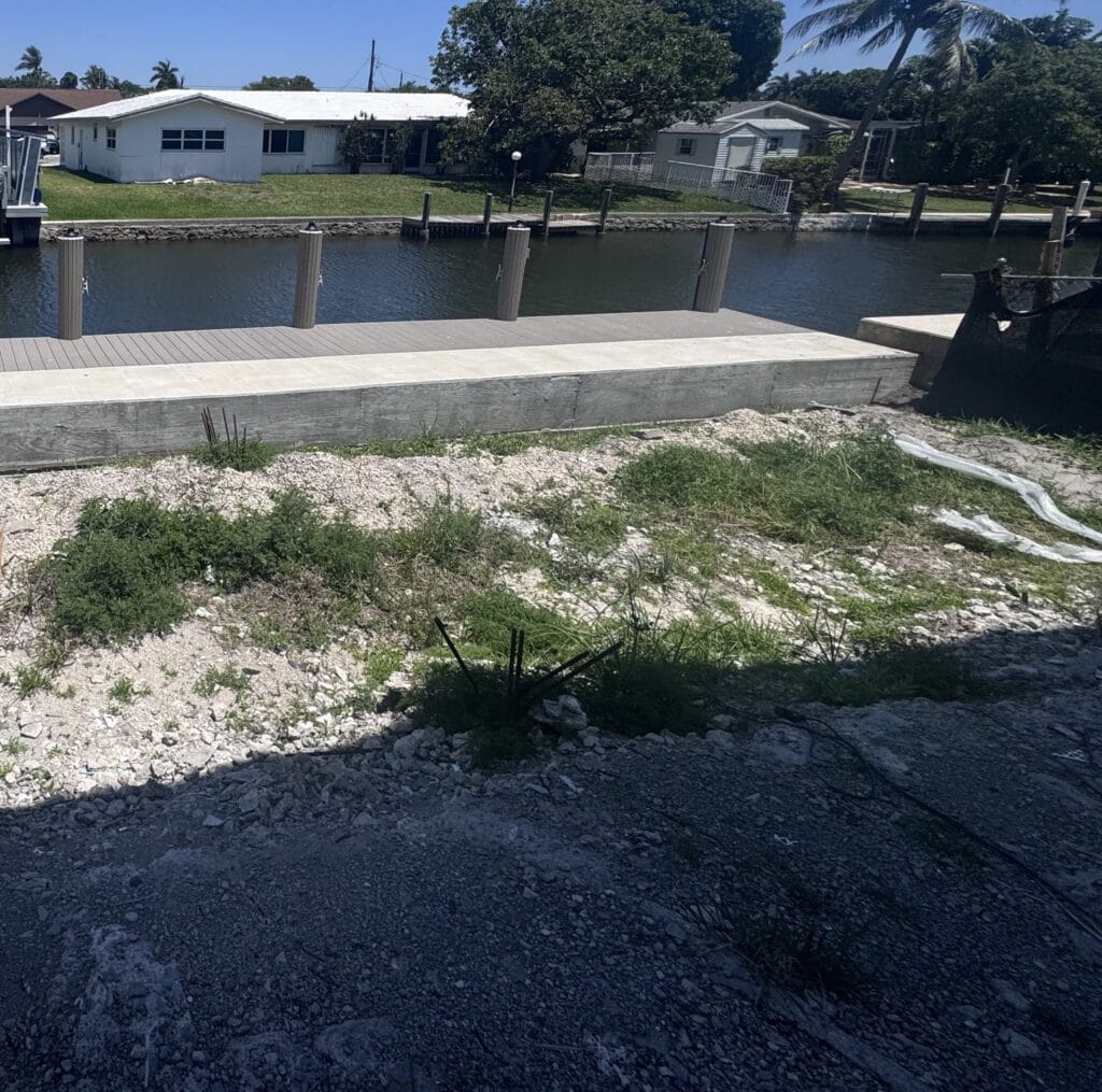 Canal frontage of the build site: new concrete seawall cap, composite dock and mooring piles, with the lot rough-graded behind it