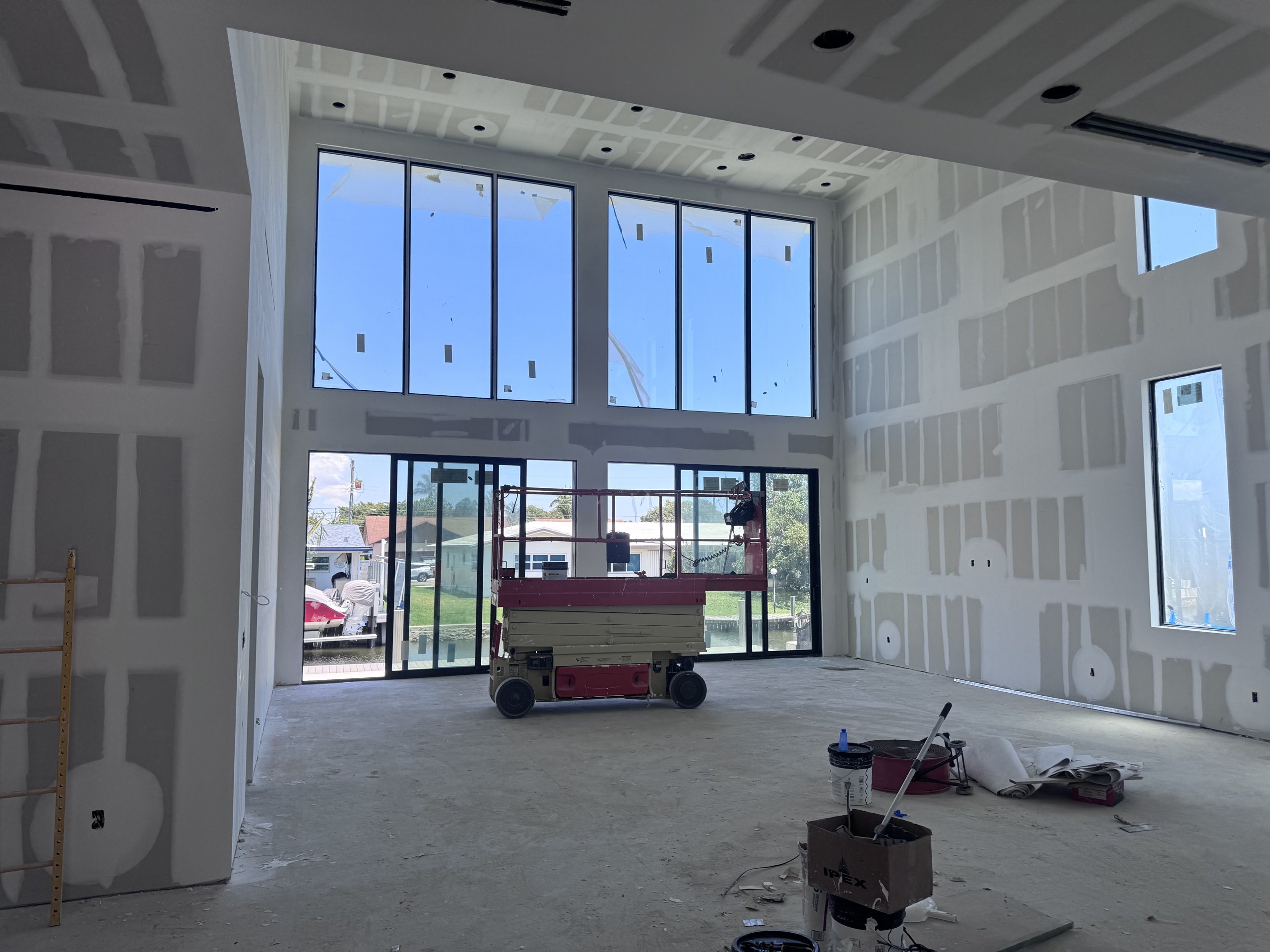 Double-height great room of an Elbaz Construction custom home at drywall stage, with a two-storey window wall and a glazed multi-slide opening onto the canal