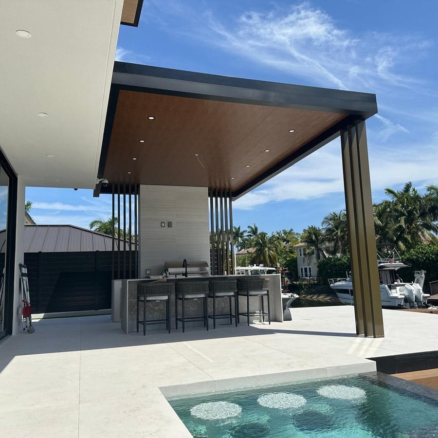 The covered terrace seen from the poolside, with the dark soffit, columns and the outdoor kitchen counter beneath it