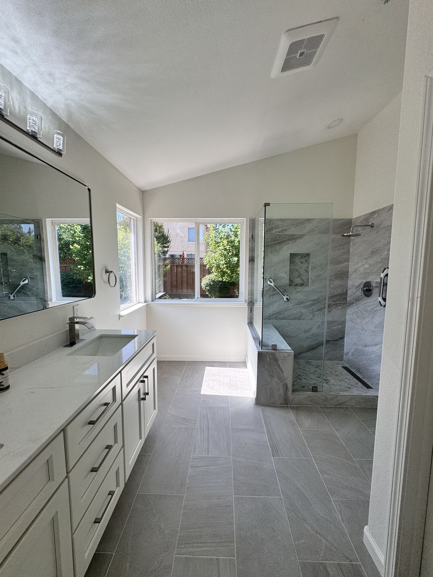 Walk-in shower behind a frameless glass panel at the end of a primary bathroom, double vanity to the left