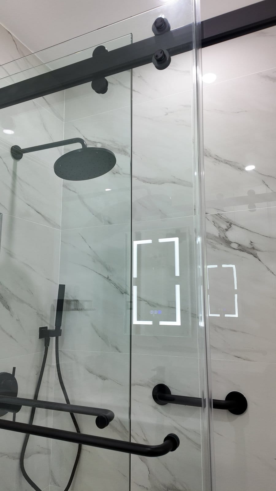 Walk-in shower behind sliding frameless glass on a black top track, with a black grab bar and rain head