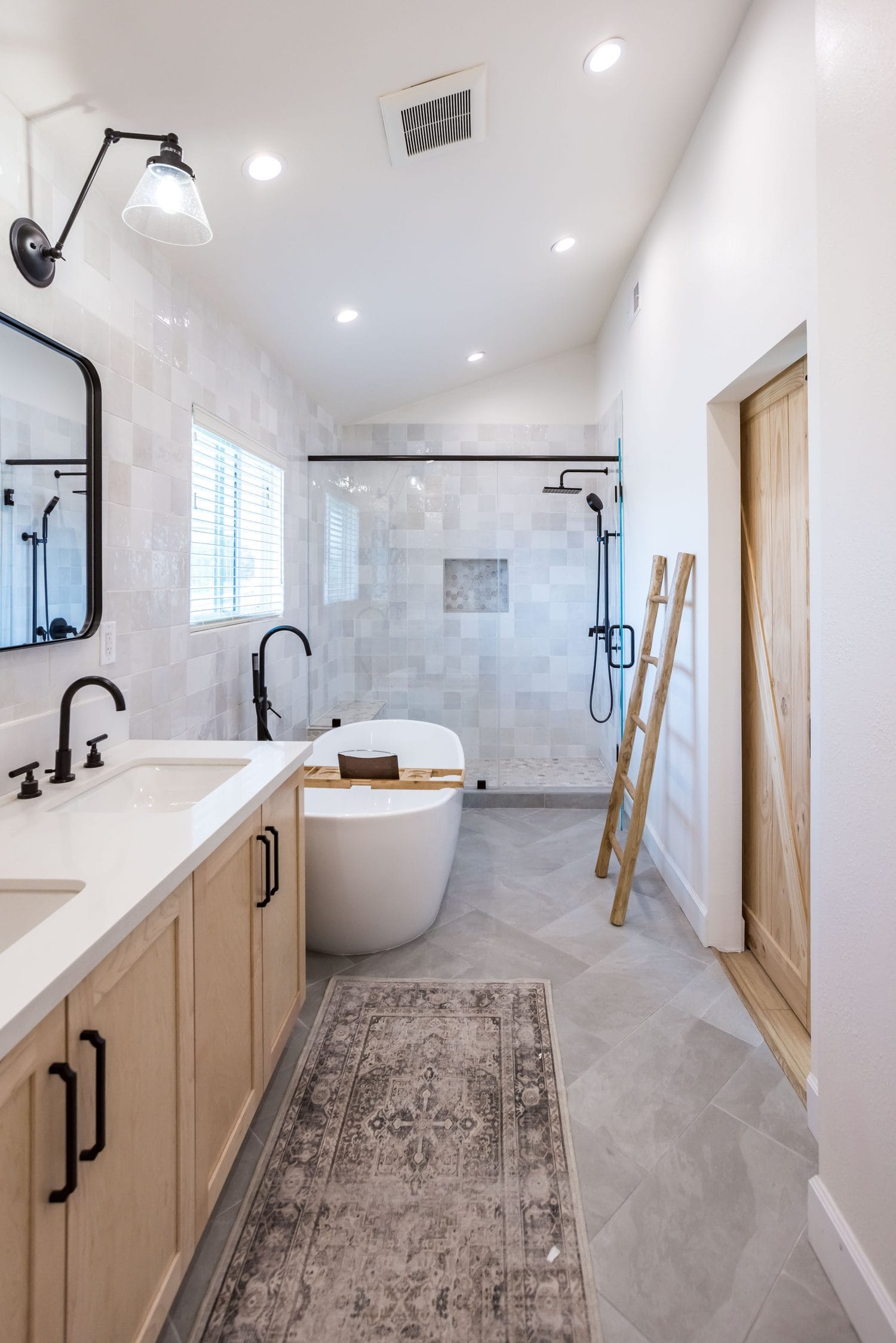 Long primary bathroom with a light oak vanity, freestanding tub and a glazed walk-in shower at the far end