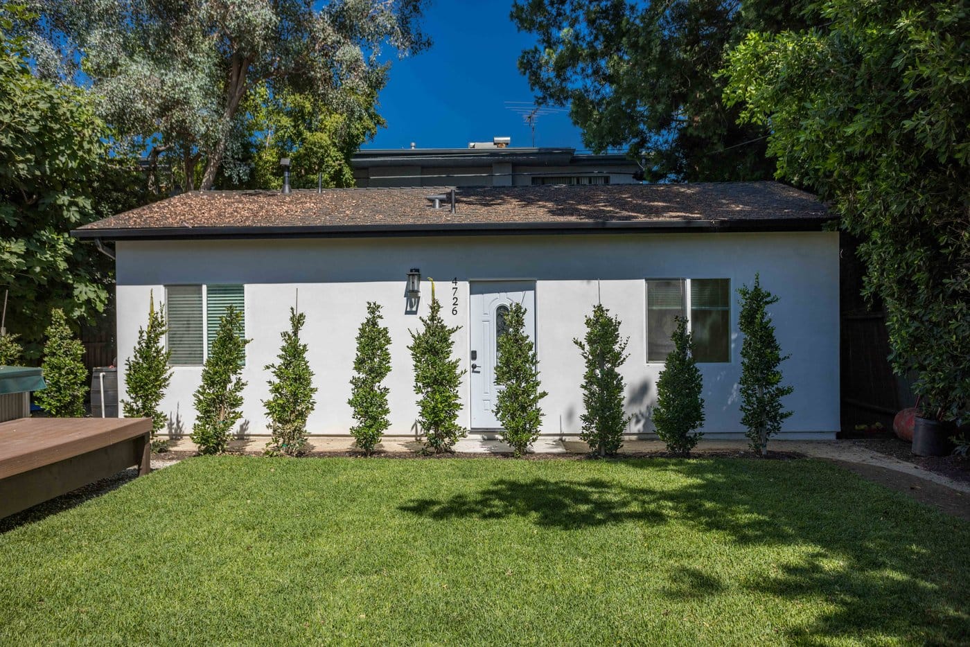 Detached converted unit with light stucco walls and a dark fascia, seen across a lawn behind a row of newly planted columnar shrubs