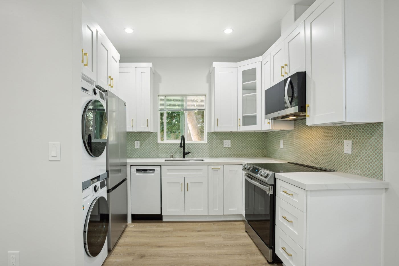 Compact white shaker kitchen with a stacked washer and dryer column, stainless range and microwave, and a green tiled backsplash