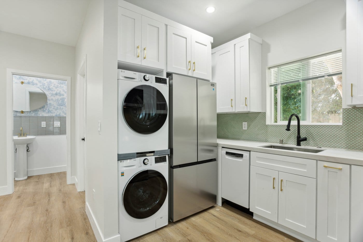 Kitchen run with a window above the sink, black tapware, green tiled backsplash and a stacked laundry column beside the refrigerator