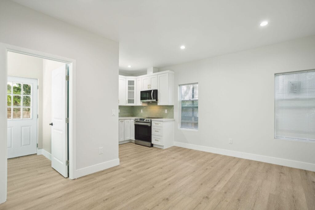 Open living area with wood-look plank flooring and a vaulted ceiling, looking through to a white kitchen at the far end