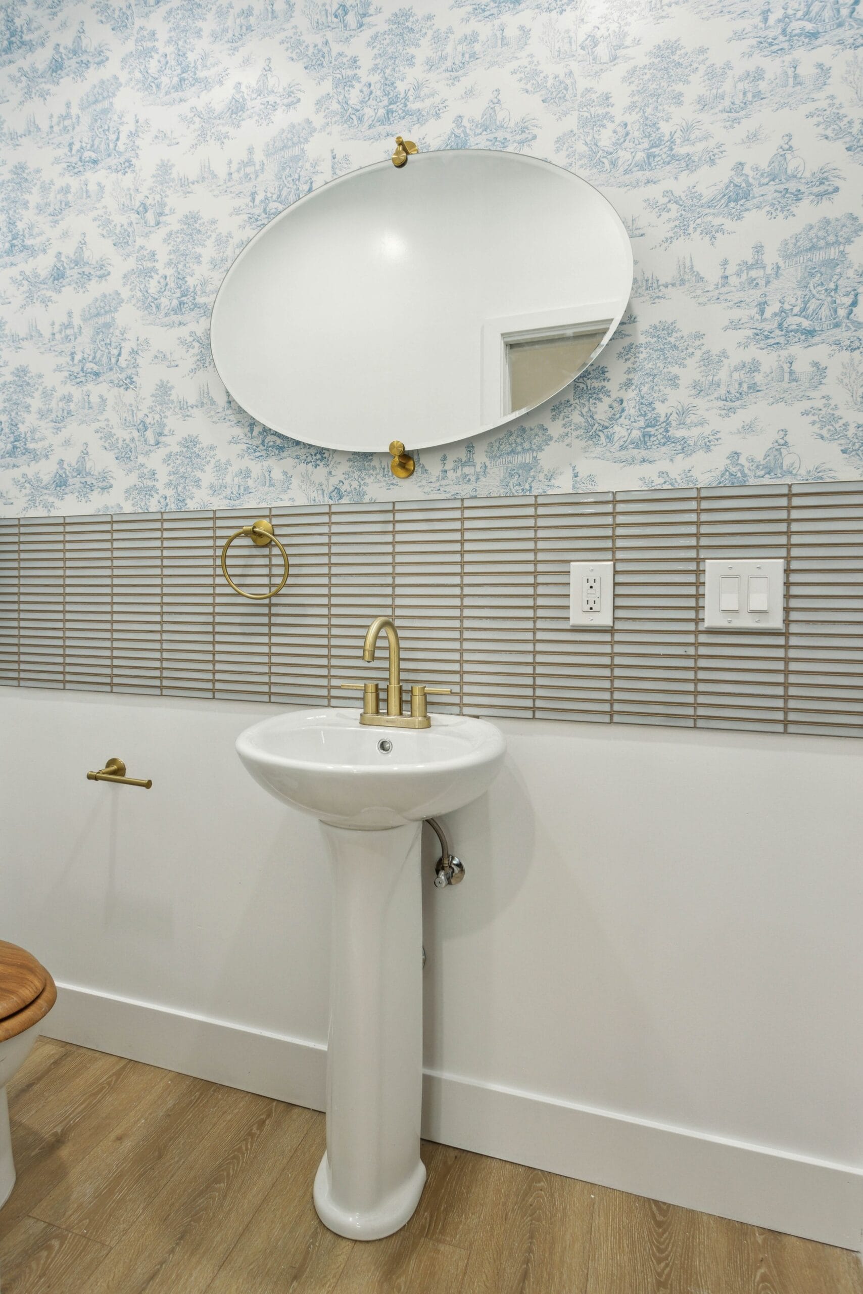 Pedestal basin with brass tapware below a round mirror, against ribbed tile and patterned wallpaper