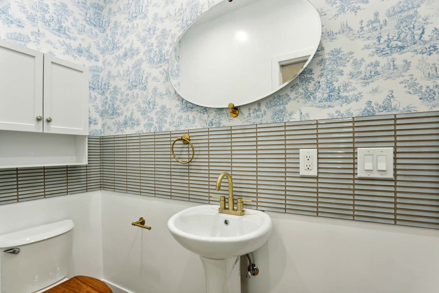 Powder room with patterned blue toile wallpaper above a ribbed tile wainscot, a round mirror and brass tapware over a pedestal basin
