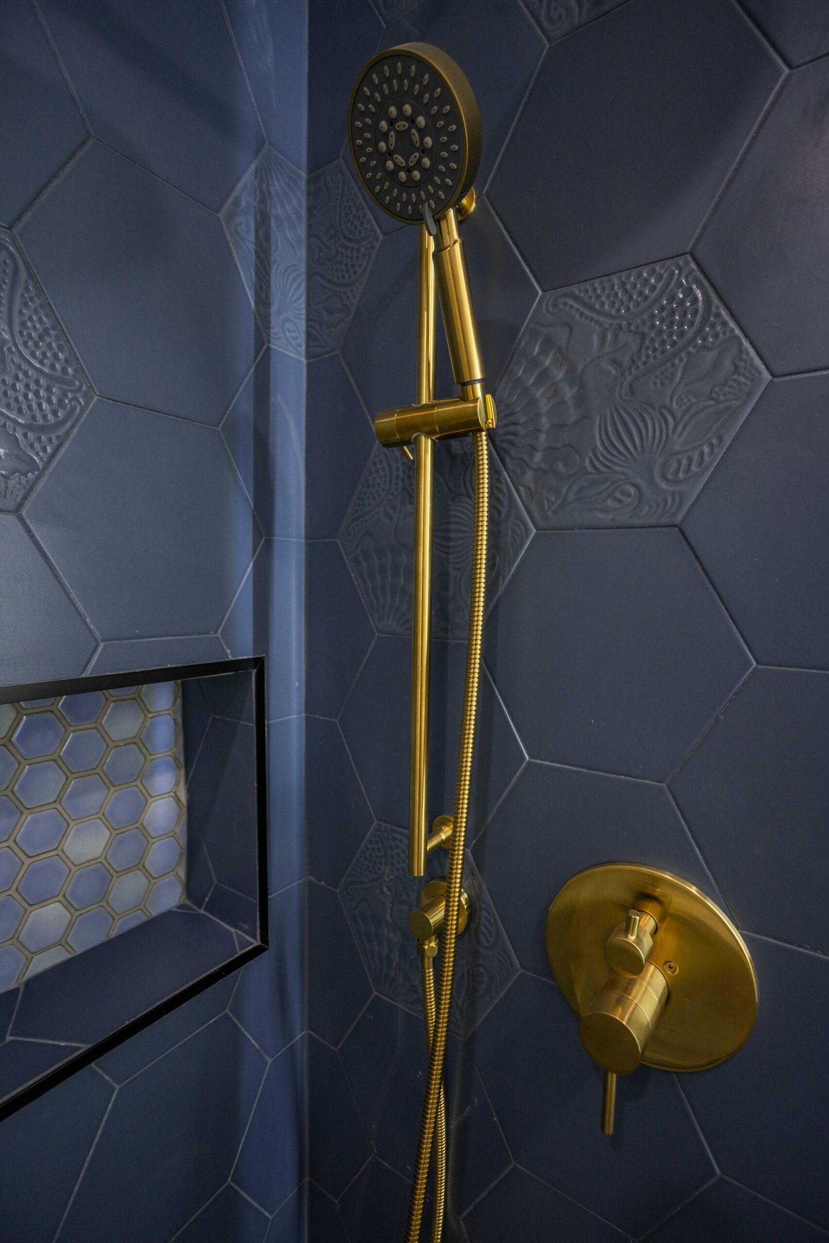 Brass shower valve and handheld on a slide bar against navy hexagonal tile beside a mosaic-lined niche
