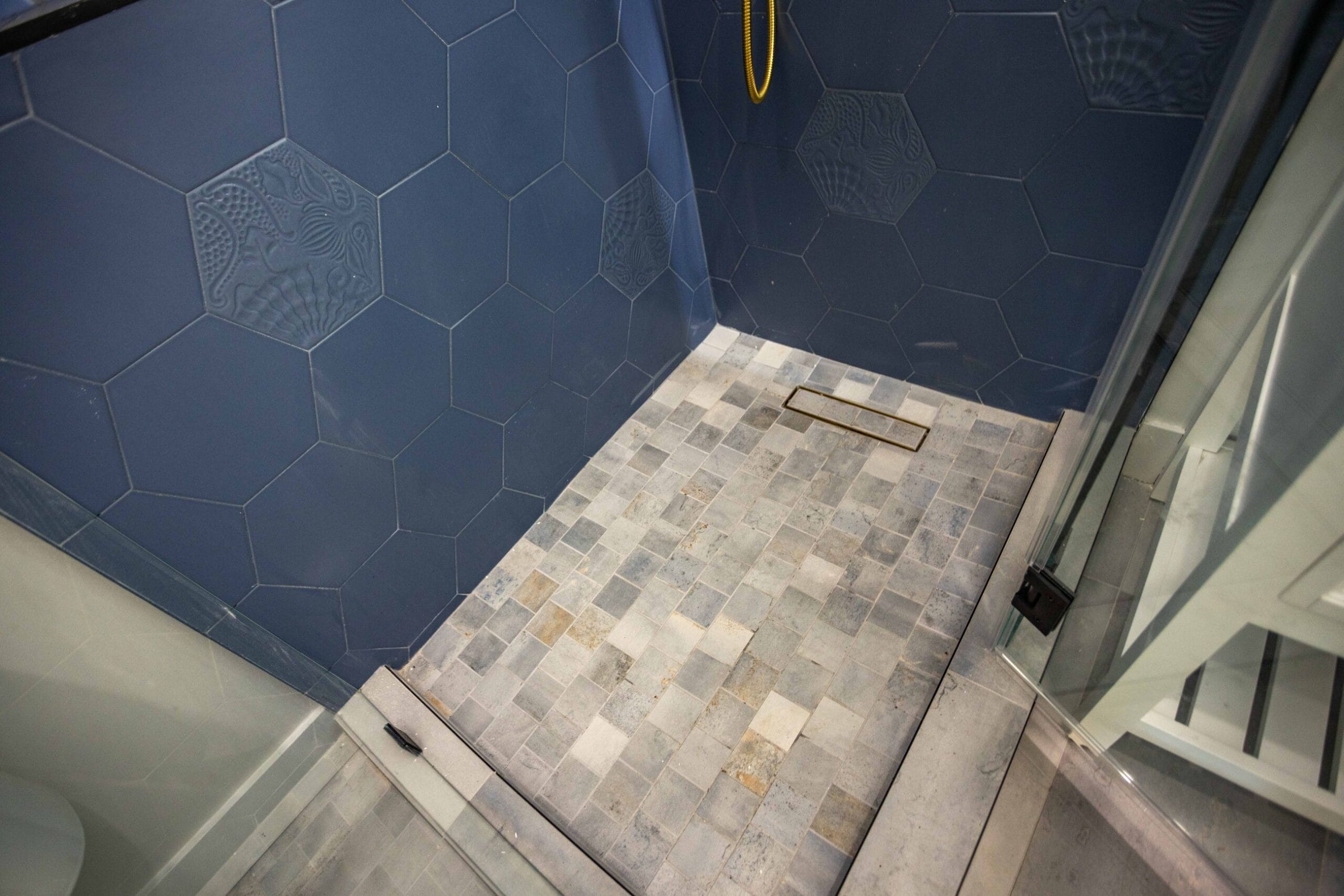 Shower floor in banded stone mosaic meeting navy hexagonal wall tile at a linear drain