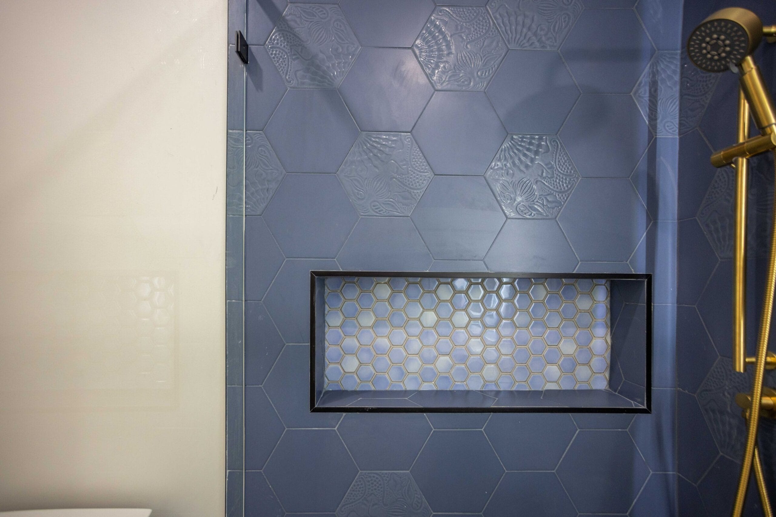Recessed shower niche lined with pale mosaic set into navy hexagonal wall tile, beside a brass handheld on a slide bar