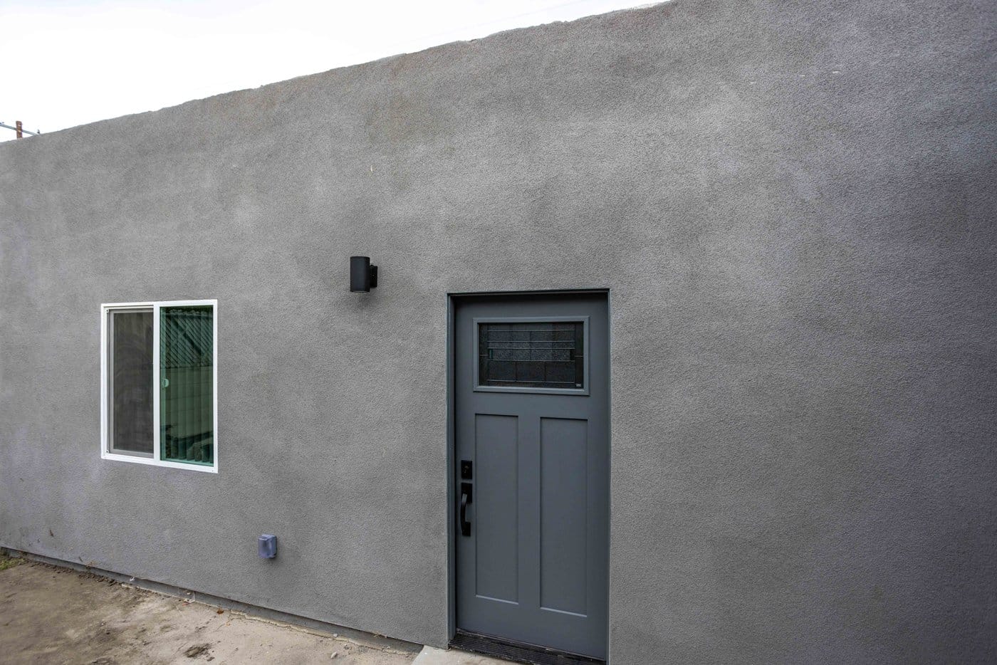 Converted garage with grey stucco walls, a flat parapet roofline, a white-framed window and a panelled entry door with a glazed insert
