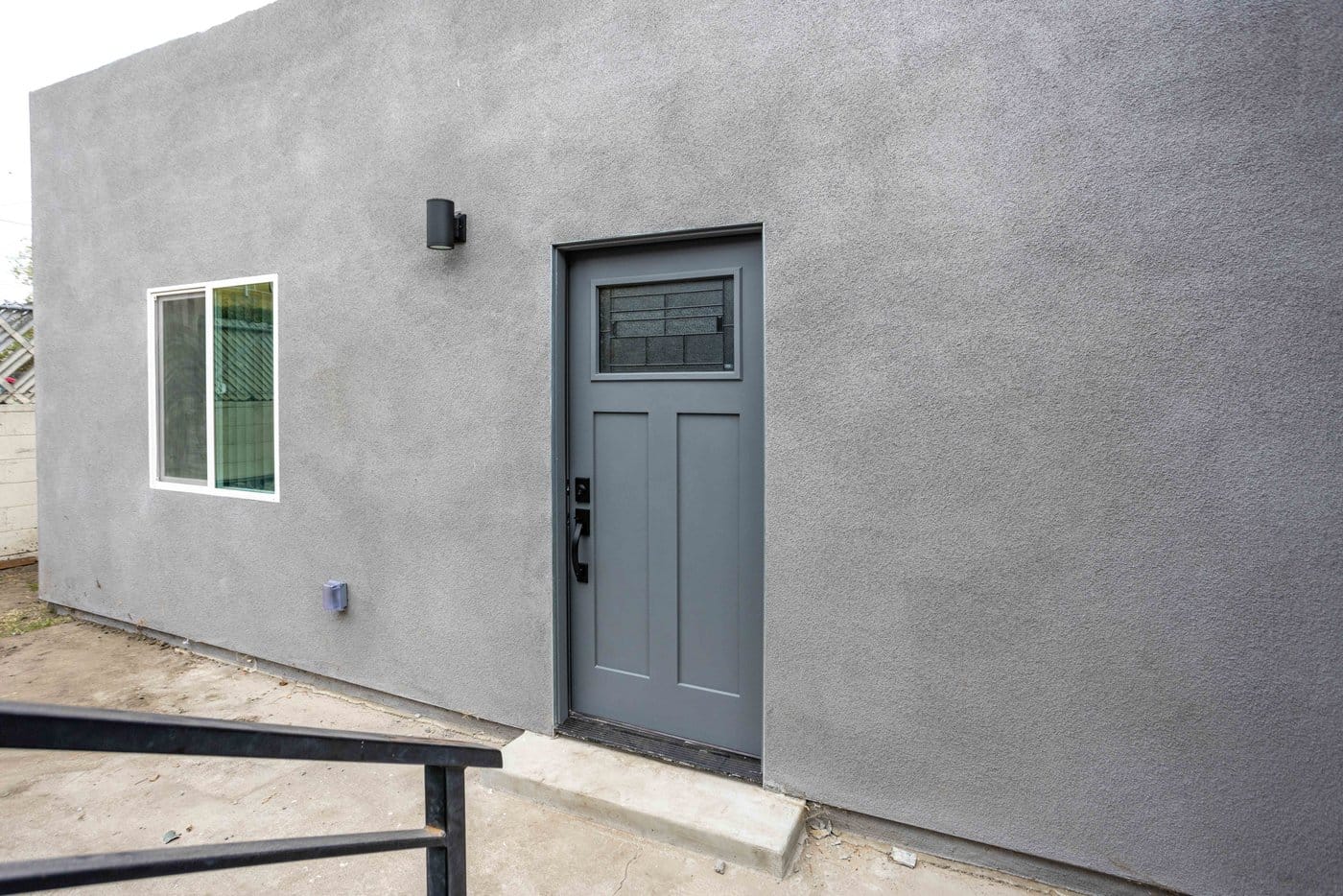 Entry door of a converted garage with a wall sconce, a black steel handrail and a poured concrete step