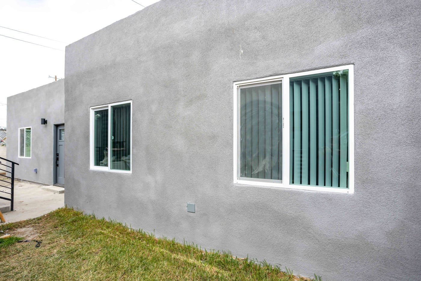 Grey stucco elevation of a converted garage with two white-framed windows above a lawn and concrete walkway