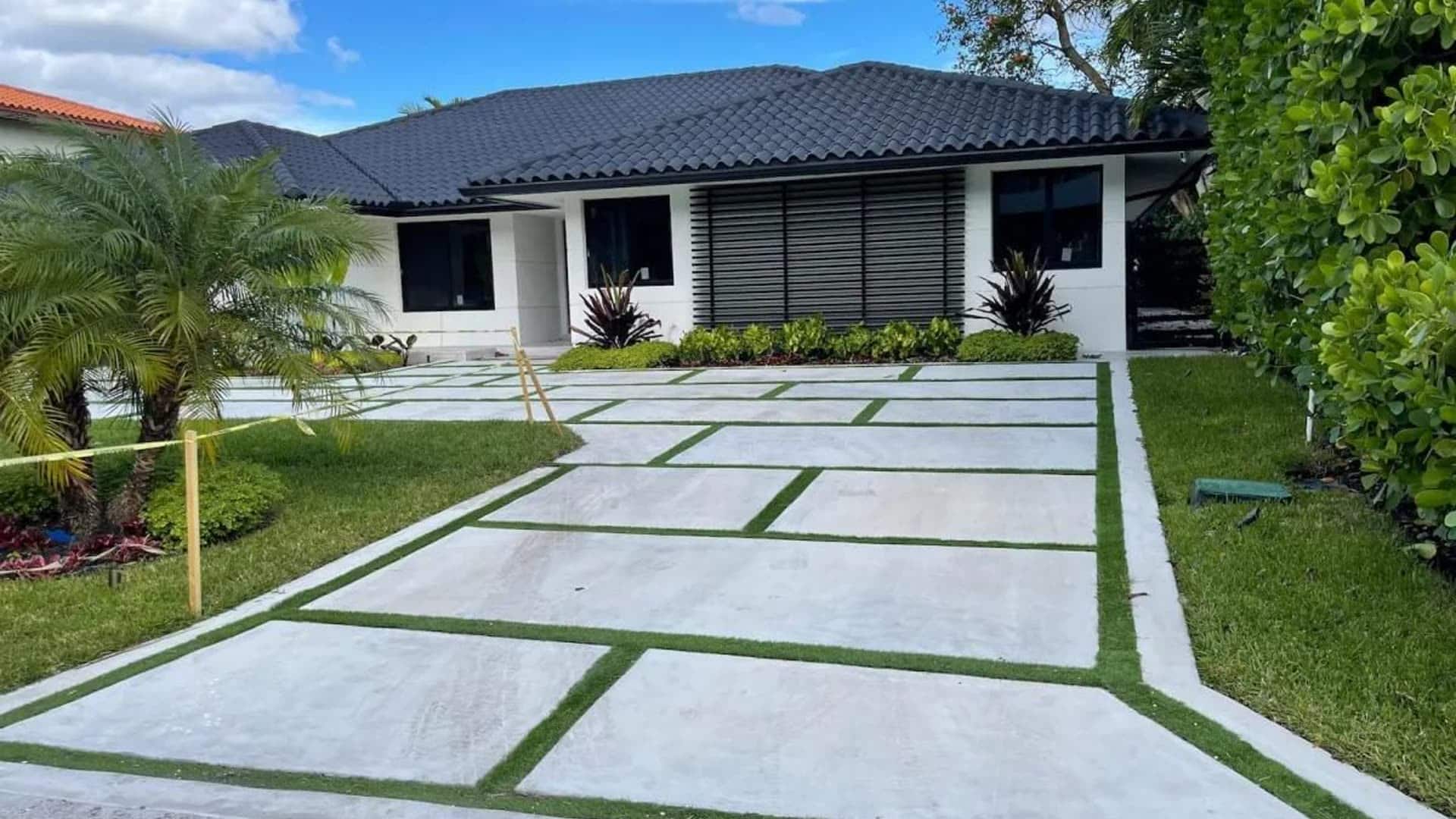 Newly finished concrete driveway with planted turf joints running to a single-storey South Florida house, still taped off after the pour