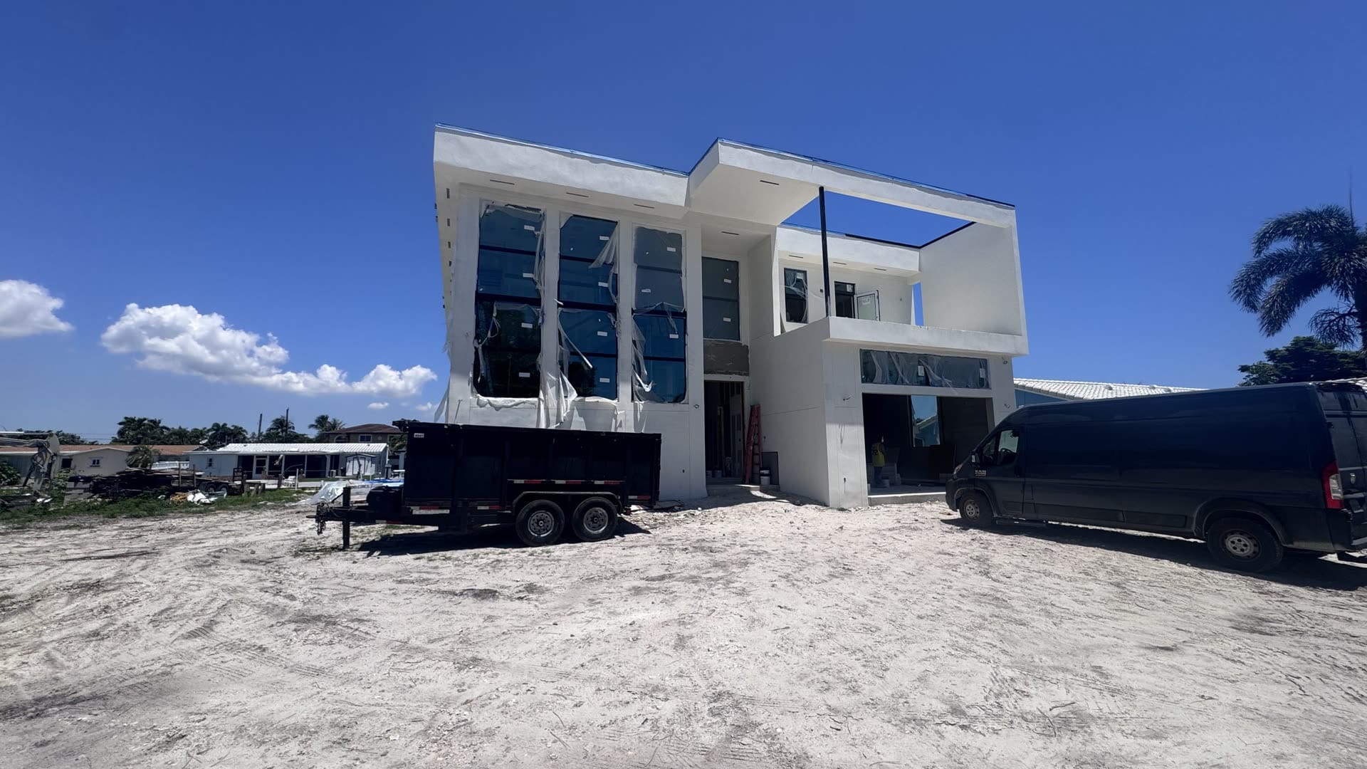 Two-storey modern custom home under construction on a cleared South Florida lot, stucco complete and glazing installed with protective film still in place