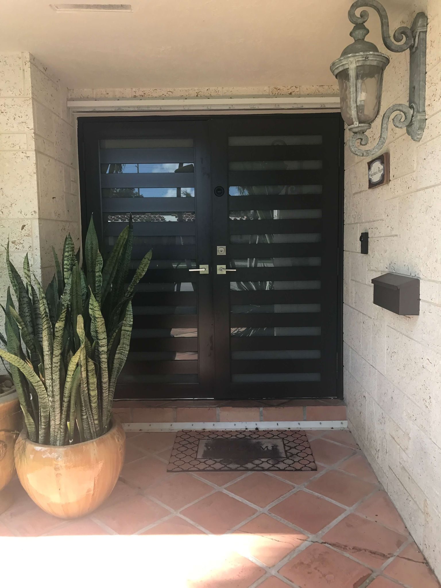Dark contemporary entry door with horizontal slatted glazing, a wall lantern alongside and terracotta paving at the threshold