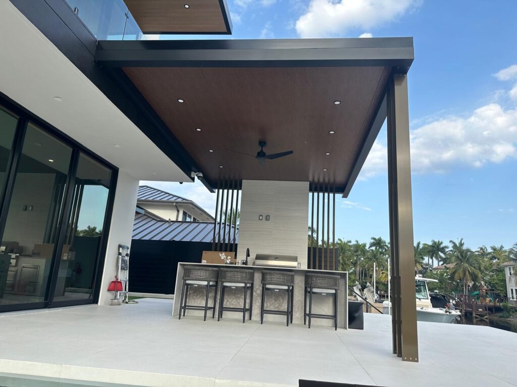 Outdoor kitchen counter with bar stools under a slatted timber ceiling, opening onto a waterway