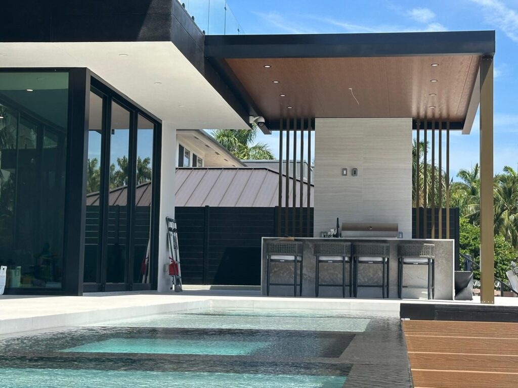 Built-in grill and stainless cabinet run set into a masonry base beneath a covered structure, with a pool and spa in the foreground
