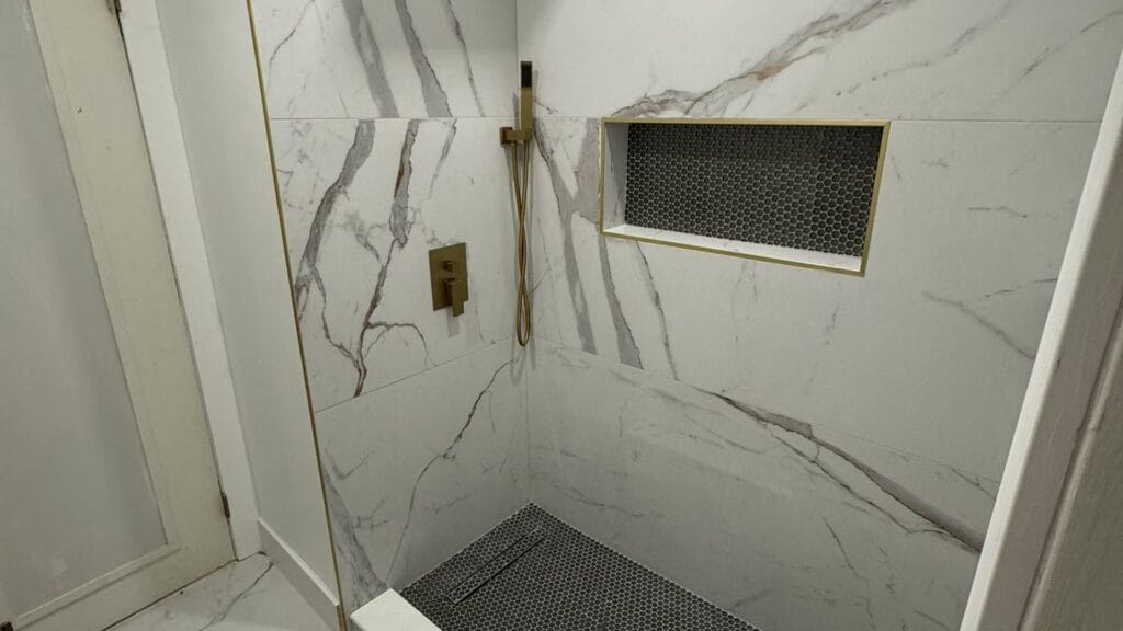 Walk-in shower finished in large-format marble-look porcelain, with a recessed niche, a linear drain at the back wall and a fixed glass panel