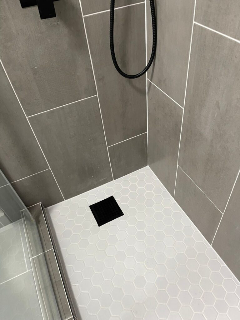 Shower floor tiled in pale hexagonal mosaic with a square centre drain and a matt black trim, seen from the opening