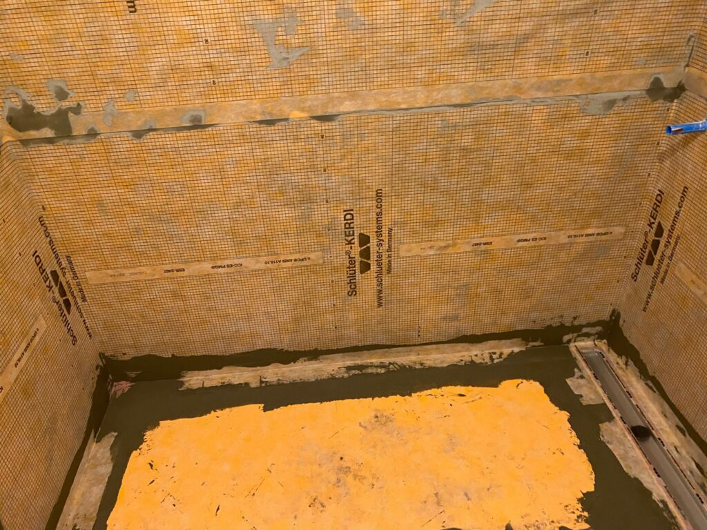Shower alcove waterproofed with a bonded orange membrane over the walls and floor, with the linear drain body set and the sloped bed formed