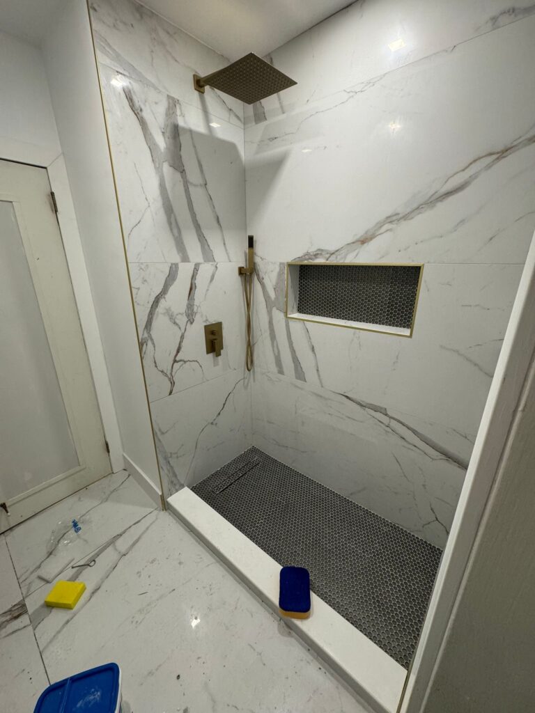 Walk-in shower finished in large-format marble-look porcelain, with a recessed niche, a linear drain at the back wall and a fixed glass panel