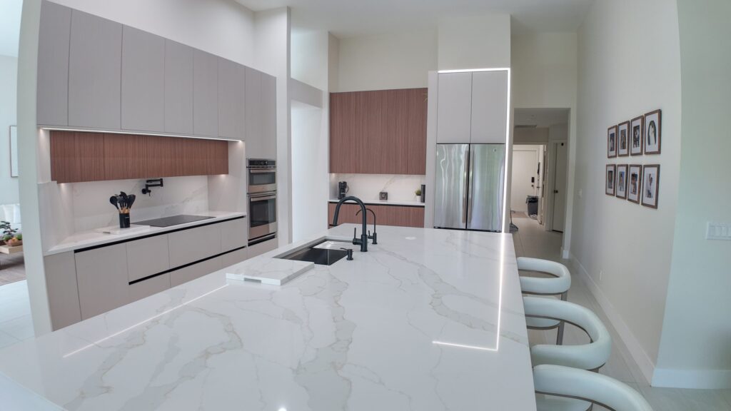 Open-plan kitchen and living remodel by Elbaz Construction in South Florida: handleless cabinetry, walnut wall units, large-format porcelain worktops and a screened terrace beyond the glazing