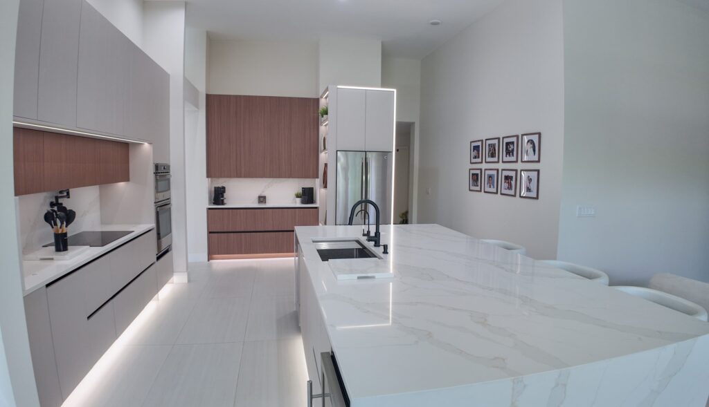 Open-plan kitchen and living remodel by Elbaz Construction in South Florida: handleless cabinetry, walnut wall units, large-format porcelain worktops and a screened terrace beyond the glazing