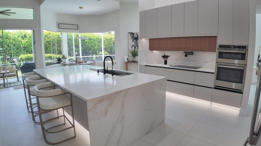 Open-plan kitchen and living remodel by Elbaz Construction in South Florida: handleless cabinetry, walnut wall units, large-format porcelain worktops and a screened terrace beyond the glazing