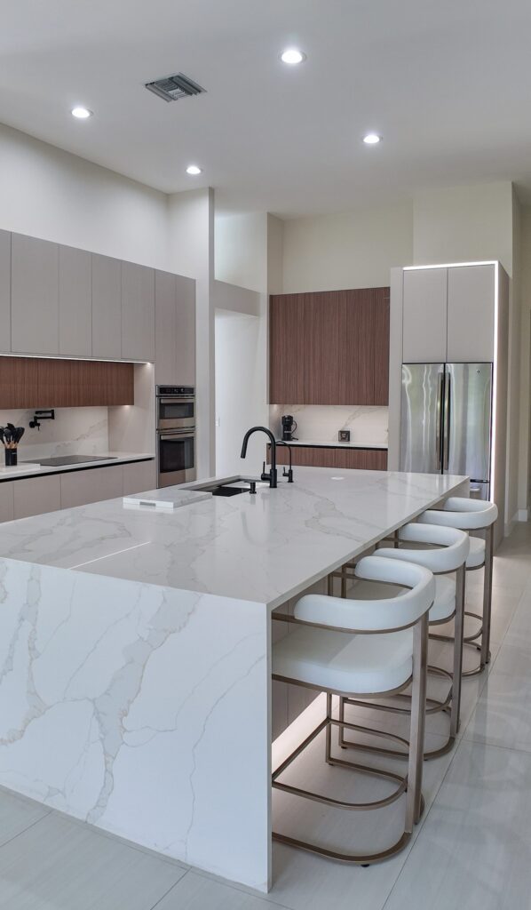 Open-plan kitchen and living remodel by Elbaz Construction in South Florida: handleless cabinetry, walnut wall units, large-format porcelain worktops and a screened terrace beyond the glazing