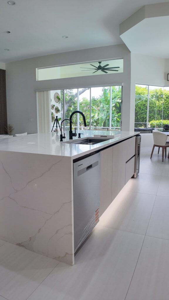 Open-plan kitchen and living remodel by Elbaz Construction in South Florida: handleless cabinetry, walnut wall units, large-format porcelain worktops and a screened terrace beyond the glazing