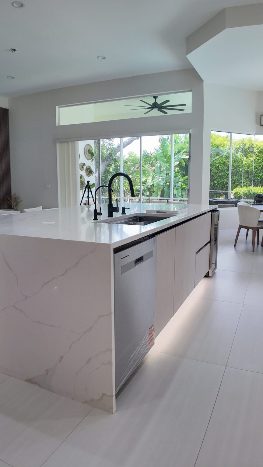 Open-plan kitchen and living remodel by Elbaz Construction in South Florida: handleless cabinetry, walnut wall units, large-format porcelain worktops and a screened terrace beyond the glazing