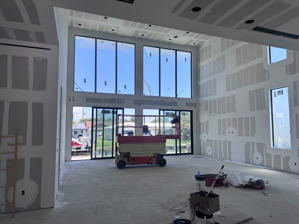 Interior of a custom home under construction by Elbaz Construction in South Florida: double-height glazing, a steel stair under installation and drywall at taping stage