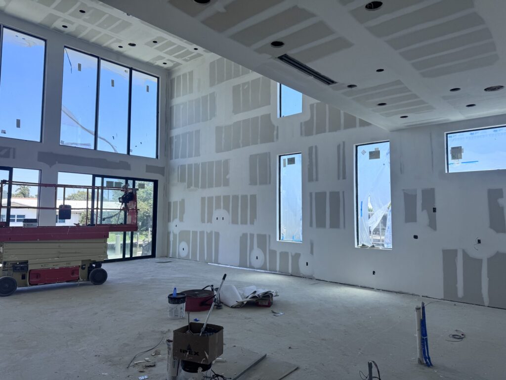Interior of a custom home under construction by Elbaz Construction in South Florida: double-height glazing, a steel stair under installation and drywall at taping stage