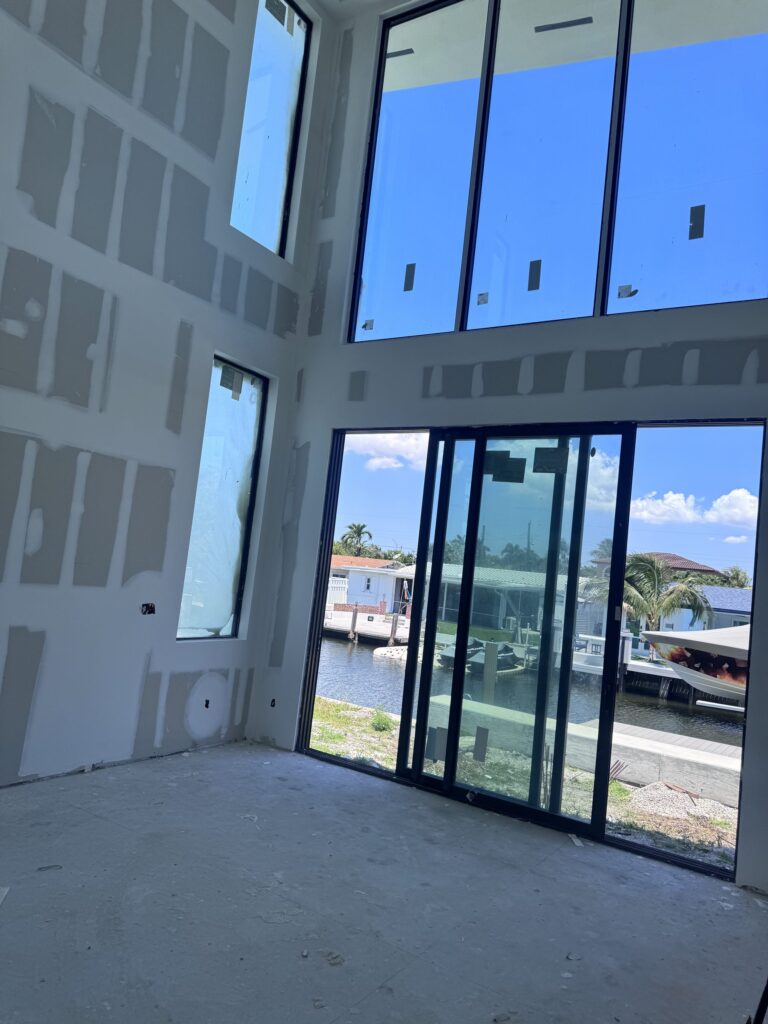 Interior of a custom home under construction by Elbaz Construction in South Florida: double-height glazing, a steel stair under installation and drywall at taping stage
