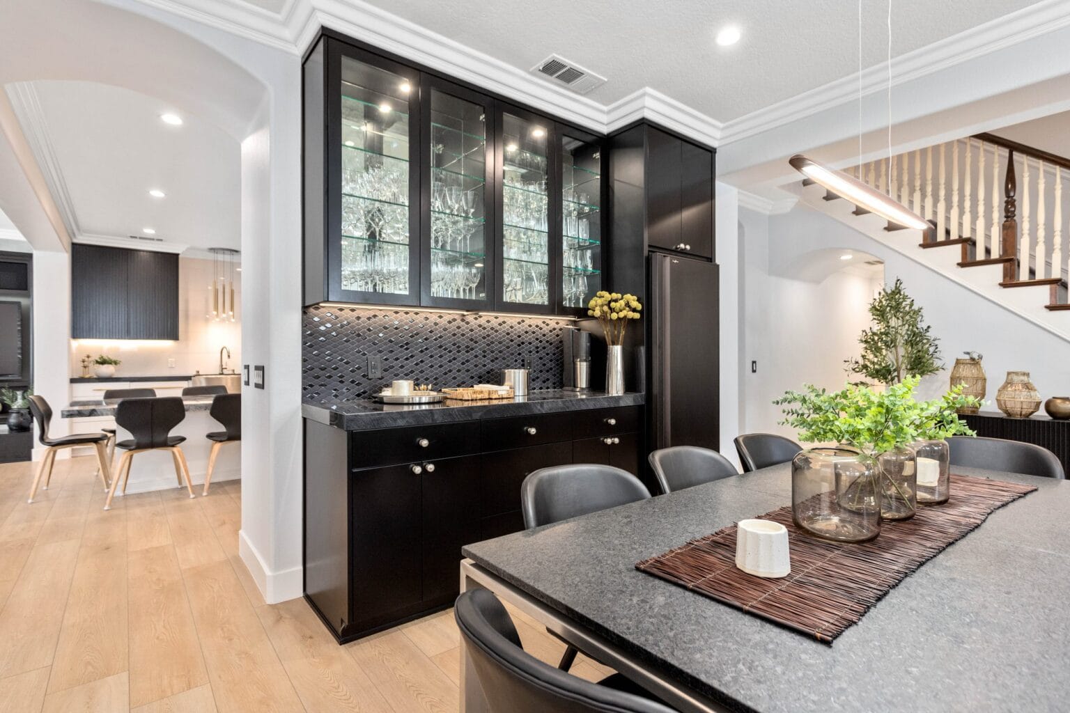 Dining table with a grey stone top in the foreground and a black servery run behind it with glass-fronted upper cabinets and a mosaic tiled splashback