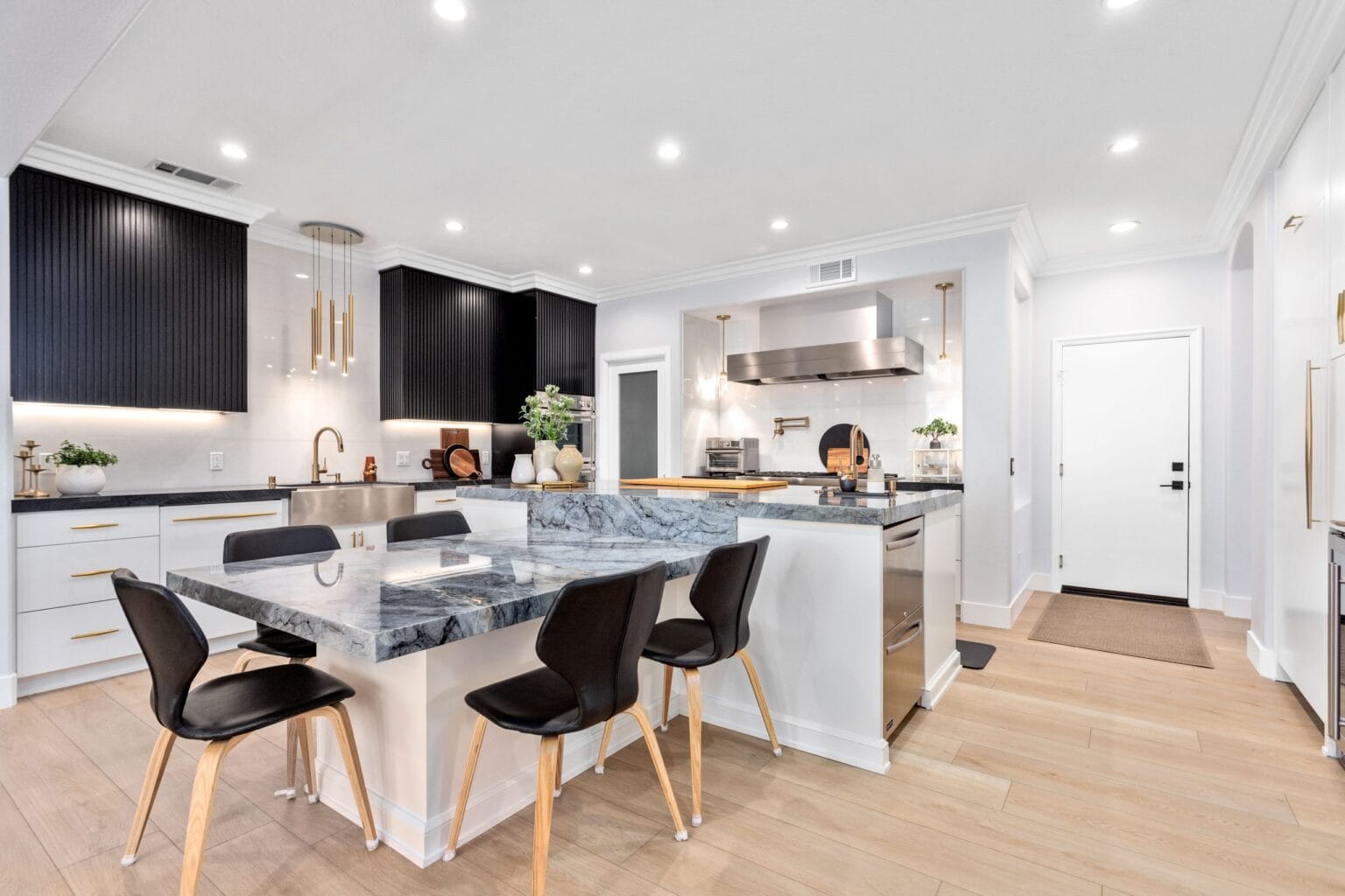 Open-plan kitchen by Elbaz Construction with a long quartzite-topped island, black dining chairs, white base cabinetry and a stainless extractor hood over the range