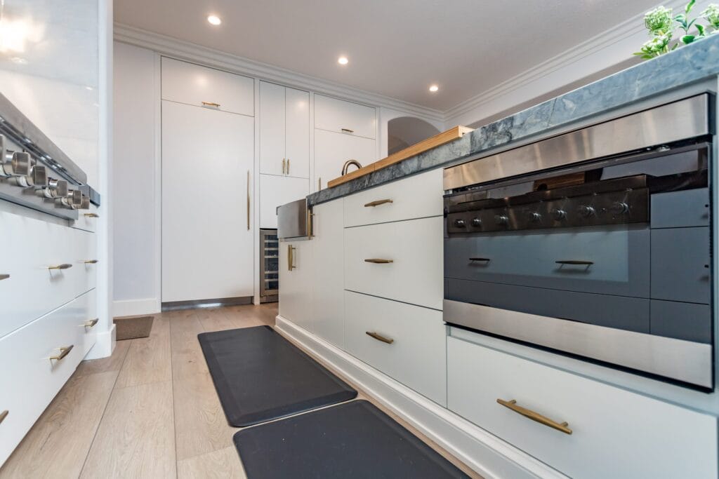 Stacked wall ovens in black glass built into a white handleless run with brushed-brass handles, on an Elbaz Construction kitchen
