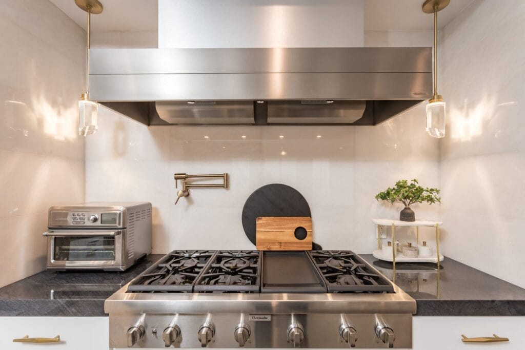 Range wall close view: a professional stainless range under a stainless extractor hood, with a brushed-brass pot filler and a wall oven alongside, by Elbaz Construction