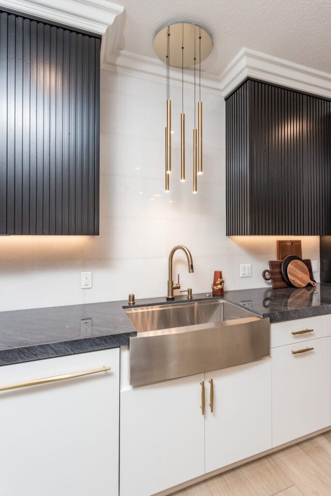 Stainless apron-front sink with a brushed-brass tap below a black fluted panel and brass pendant lights, on an Elbaz Construction kitchen remodel