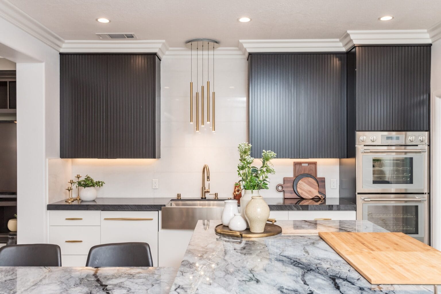The kitchen from the sink side, with brass pendants, built-in wall ovens and a timber board on the quartzite island