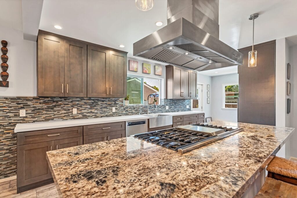 Granite-topped island with a gas hob below a stainless island extractor, in a grey shaker kitchen by Elbaz Construction