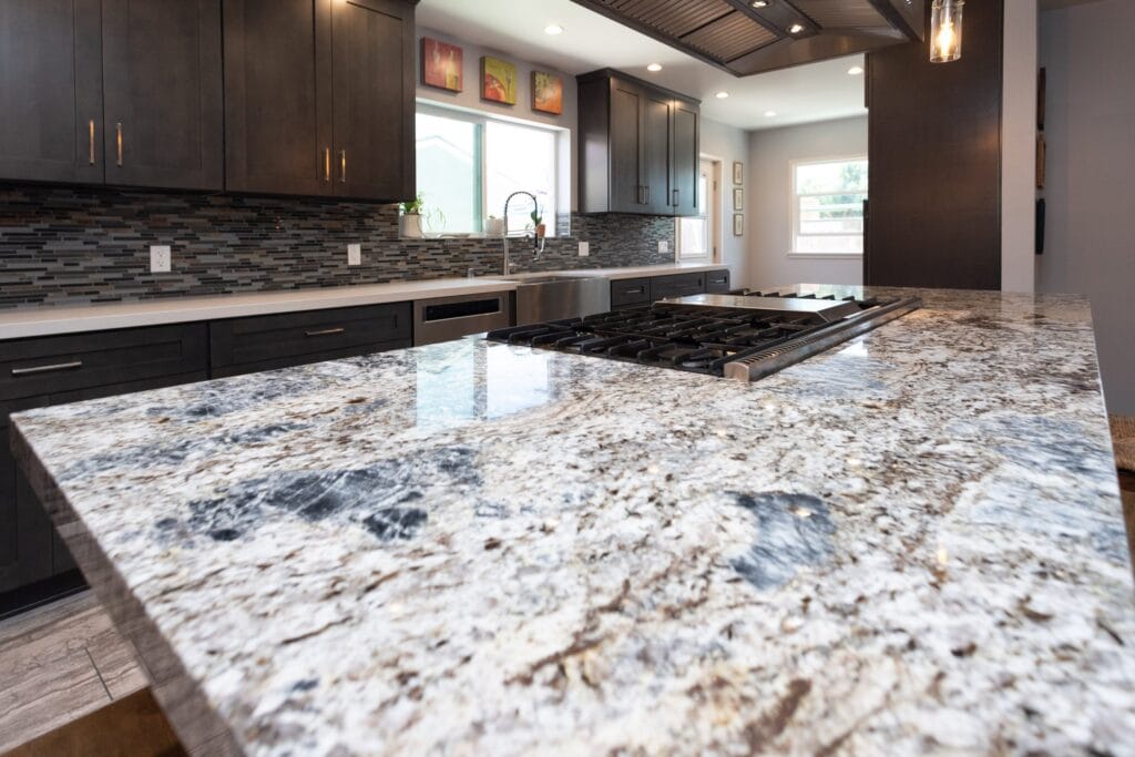 Close view along a heavily figured granite island worktop to the gas hob and the mosaic splashback beyond, by Elbaz Construction
