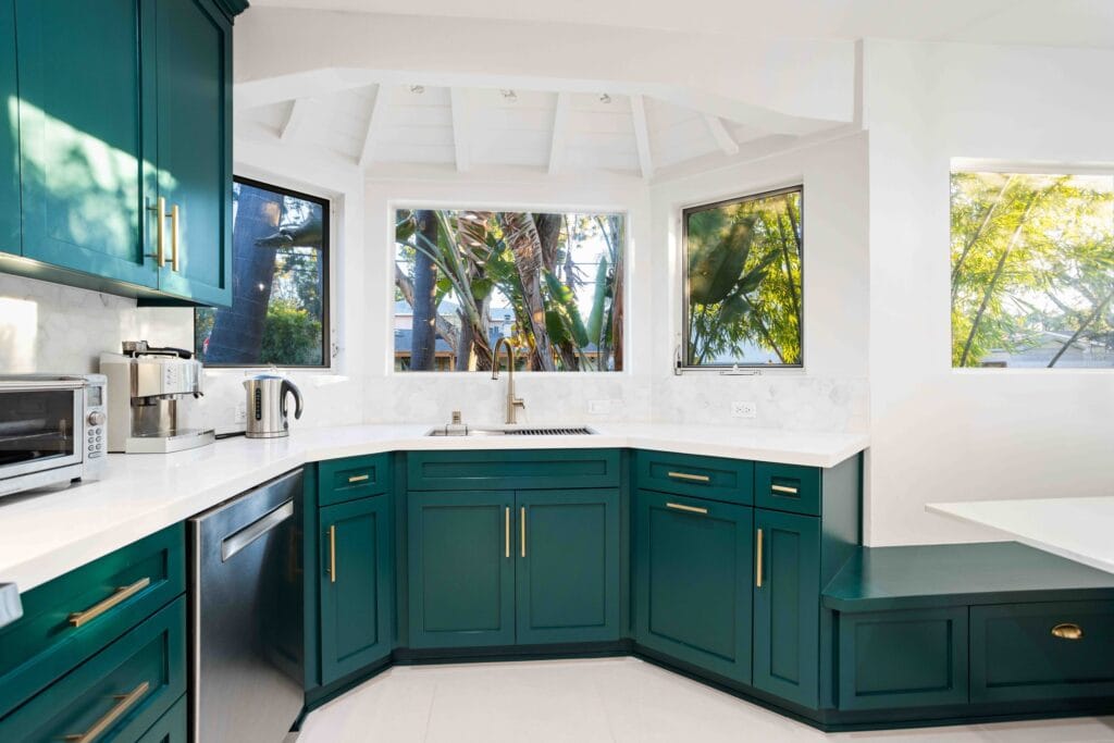 Corner run of teal shaker units with brass handles under a bank of windows, on an Elbaz Construction kitchen remodel