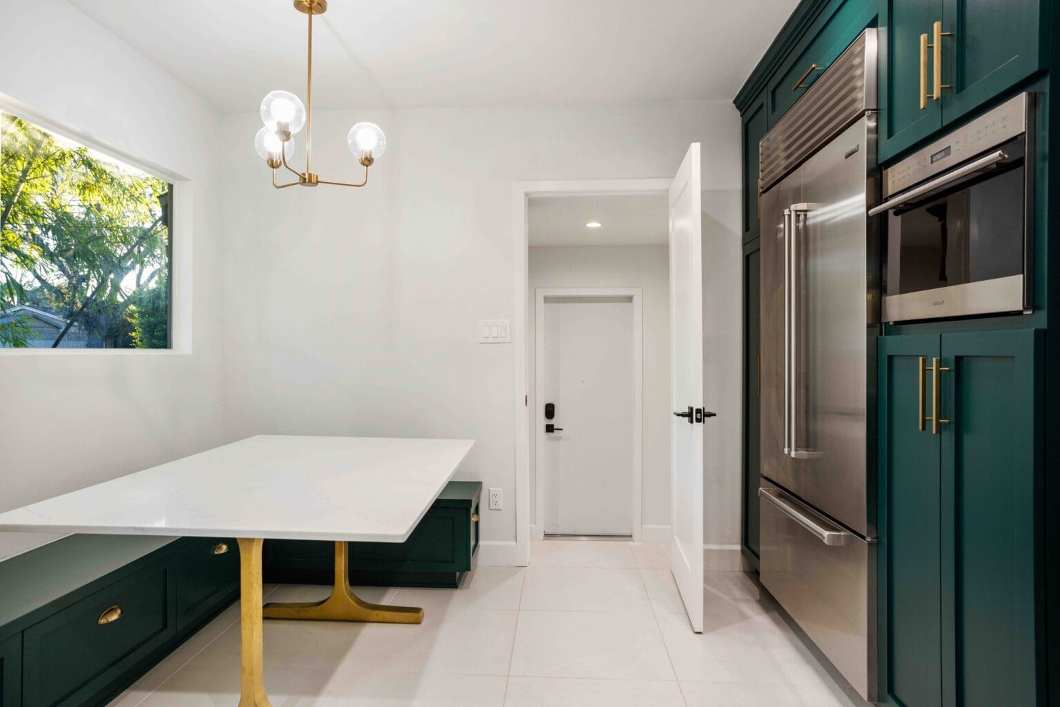 Teal shaker kitchen by Elbaz Construction with a quartz-topped banquette table on a brass pedestal, a built-in fridge and a wall oven column beyond