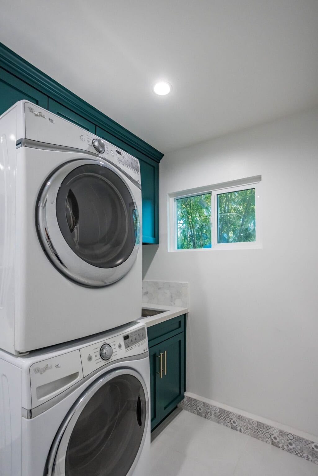 Utility room with a stacked washer and dryer, a teal base cabinet and a small window, finished by Elbaz Construction