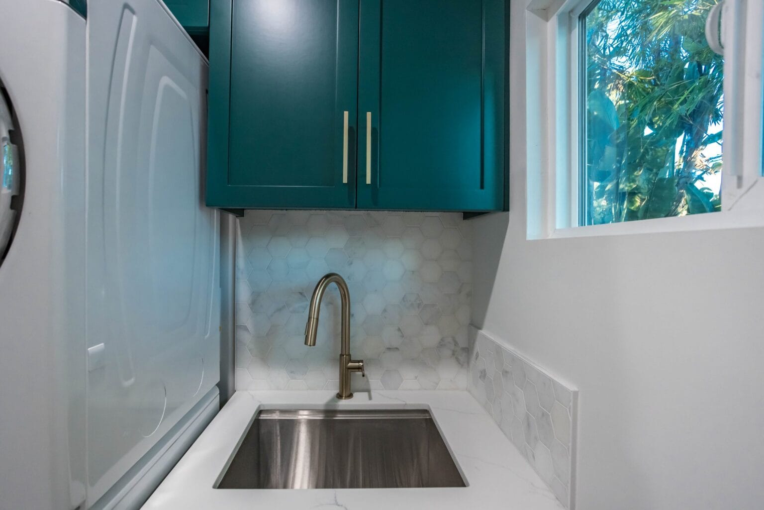 Utility room sink set in a white worktop below a window, with teal shaker upper cabinets and a hexagonal marble splashback