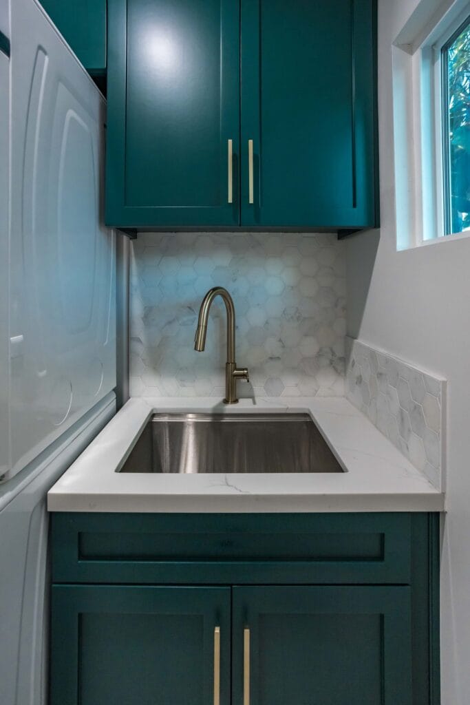 Close view of the utility sink, brass tap and hexagonal mosaic splashback below a teal wall cabinet, by Elbaz Construction